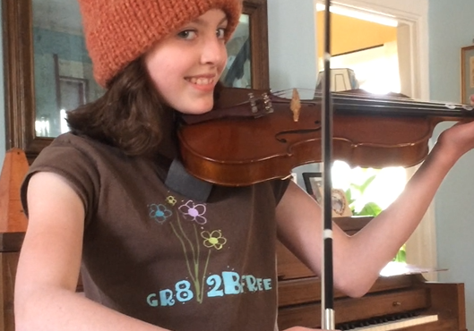 Young girl, Ella Thompson Rose playing the violin as she smiles into the camera, wearing a burnt orange knit hat and a piano in the background.