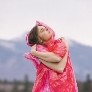 photo of Francesca Blanchard standing with her arms crossed looking up to the right. She is wearing a pink rain poncho and mountains blurry in the background.