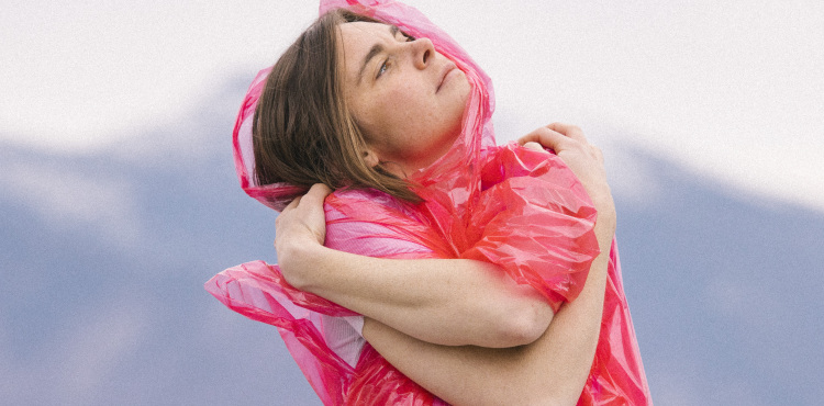 photo of Francesca Blanchard standing with her arms crossed looking up to the right. She is wearing a pink rain poncho and mountains blurry in the background.